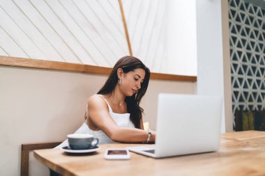 Genç İspanyol kadın serbest yazar, uzaktan çalışma sırasında not defterine not alırken elinde bir fincan kahve akıllı telefonu ve dizüstü bilgisayarla ahşap masada oturuyor.