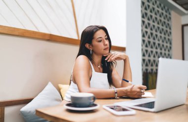 Etnik serbest çalışan kadın elinde bir fincan kahve, akıllı telefon ve dizüstü bilgisayarla masada oturuyor ve not defterinden notlar alırken ekrana bakıyor.