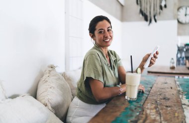 Elinde bir bardak limon dilimi ve samanla oturmuş elinde cep telefonuyla kameraya bakan pozitif, genç bir İspanyol kadın.