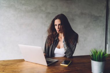 Düşünceli genç bayan serbest çalışan gündelik kıyafetlerle tahta masada dizüstü bilgisayarı ve akıllı telefonuyla uzaktan kumandalı bir proje üzerinde çalışıyor.