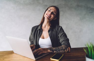 Kolunda dövmesi olan, gülümseyen, cep telefonuyla masada oturan ve kafedeki iş projesinde uzaktan çalışırken internet üzerinden yazan serbest çalışan bir kadın.