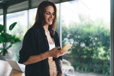Çadırda cam duvarın yanında duran, cep telefonuna bakarken masaya yaslanan ve ekrana bakan neşeli, gündelik kıyafetli genç bir kadın.