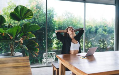 Sakin, uzun kahverengi saçlı, beyaz saçlı genç bir kadın tropik bitkinin yanındaki ahşap masada oturuyor ve elleri başının arkasında ve gözleri kapalı bir şekilde dizüstü bilgisayarla çalışırken vücudunu esnetiyor.