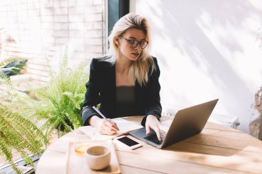 Not defteri kullanan beyaz kadın gazeteci içerik yayınlarını planlamak ve sosyal ağları taramak için dizüstü bilgisayar kullanıyor, gözlüklü yetenekli kadın netbook 'ta 4G kablosuz bağlantısına sahip.