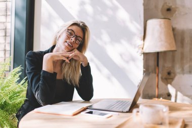 Gündüz vakti kafeteryada kameraya bakan akıllı günlük kıyafetlerle ve optik gözlüklerle başarılı kadın yöneticinin portresi, modern dizüstü bilgisayarlı yetenekli bir kadın kapalı mekanda poz veriyor.