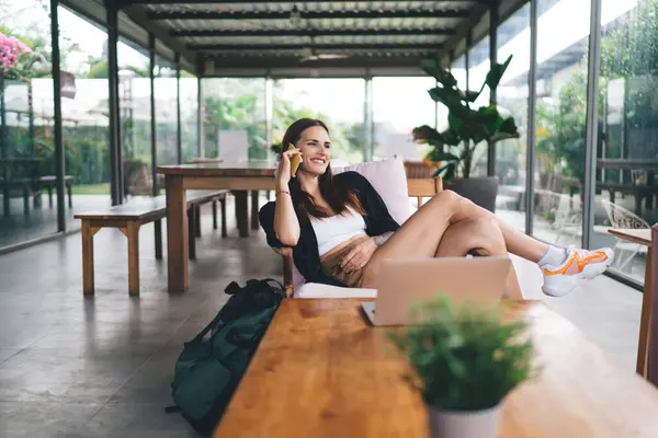 stock image Smiling young female freelancer wearing casual clothes sitting cross legged near wooden table with laptop while looking away and speaking on mobile phone