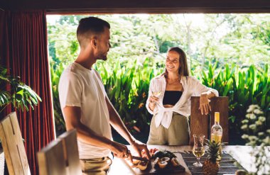 Carefree female enjoying time with Caucasian boyfriend talking and cooking tasty food at terrace, happy hipster girl holding glass of white wine smiling with her husband feeling romantic at date clipart