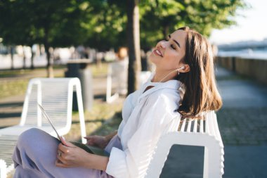 Neşeli genç bir bayanın gülümsemesi, bankta oturması, dinlenmesi ve güneşli bir havada dizüstü bilgisayardan gelen kulaklıklarla müzik dinlemesi.