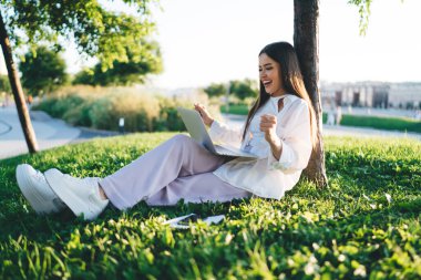 Beyaz elbiseli genç bir kadının yeşil çimlerde oturması, bacak bacak bacak üstüne atması yumruklar havada ve başarıyı kutlaması.