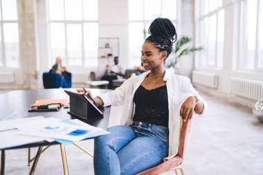 Gündelik giysiler içinde, dijital tablet kullanarak masada oturan ve yaratıcı ortak çalışma alanı projesinde çalışan pozitif etnik iş kadını.