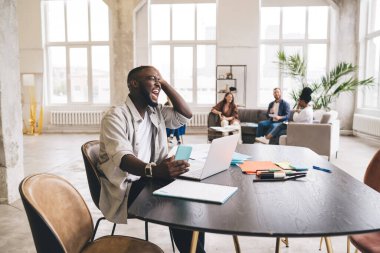 Ellerinde akıllı telefonla ahşap masada oturmuş ofiste dizüstü bilgisayar kullanarak iş projesi üzerinde çalışırken kafasına dokunup bağıran sıradan giyinen Afrikalı Amerikalı girişimci.
