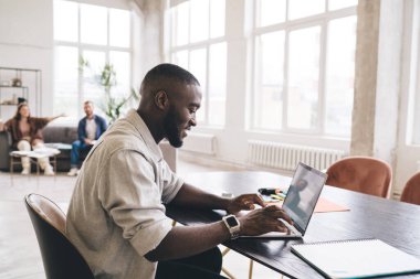 Masada oturan ve geniş bir odada dizüstü bilgisayarda çalışan Afro-Amerikan erkek çalışanın yan görüntüsü