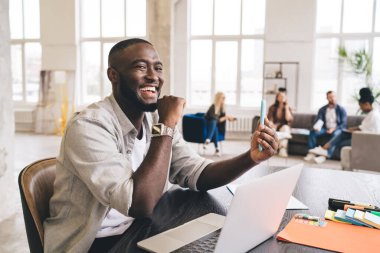 Açık tişörtlü, akıllı bir Afro-Amerikalı, arka plandaki koltuklarda oturan iş arkadaşlarıyla geniş bir odada otururken.