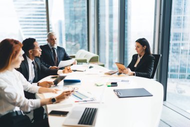 Bir grup çok ırklı iş arkadaşı resmi giysiler içinde Müdür masasında oturmuş ellerinde kağıt zımbırtılarıyla proje detaylarıyla birbirlerine bakarken iş birliği yapıyorlar.