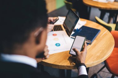 Anonim bir erkek çalışanın ahşap masada oturup, yaratıcı ofis projesinde çalışırken cep telefonuna göz atması.