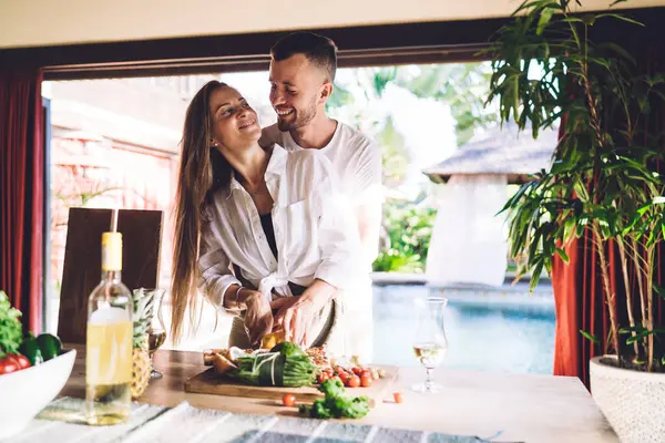 stock image Excited male and female with fresh vegetables laughing while cooking brunch meal at house terrace, joyful Caucaisan marriage with food smiling and rejoicing togetherness in homey interior