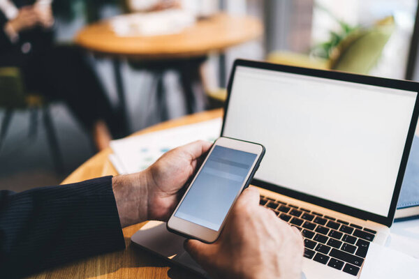 Crop anonymous male freelancer sitting at wooden table with laptop and browsing mobile phone while working on business project in office