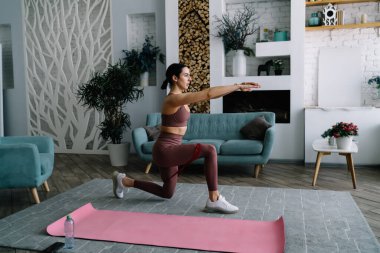 Evde sırt ağrısını azaltmak için rahat oturma odasında yoga yaparken dizlerinin üzerine çöküp kollarını öne uzatan genç bir kadının yan görüntüsü.