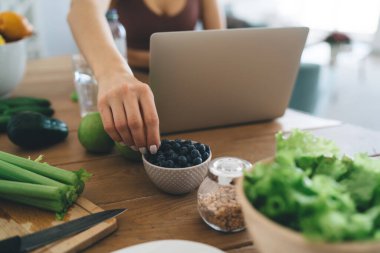 Portatif bilgisayarda video tarifi izleyen ekin dişisi masada oturup mutfakta sağlıklı yiyecekler hazırlarken kaseden böğürtlen alıyor.