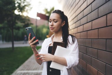 Kitap çantalı, gülümseyen ve cep telefonlu genç bir kız öğrenci ekrana bakıyor ve tuğla duvar binasının yanında duruyor.