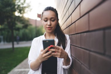 Kampüsteki tuğla duvara yaslanıp mesaj gönderirken beyaz tişörtlü, cep telefonu ekranına bakan pozitif genç bir kadın.