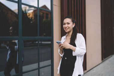 Modern ofis binasının cam penceresinin yanında duran müziği dinlerken akıllı telefon sörfü yapan pozitif bir kadının yansıması.