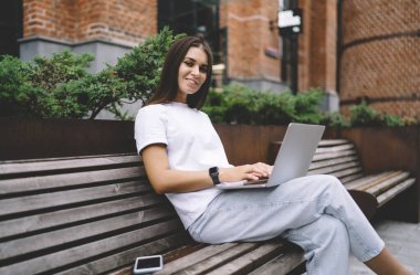 Laptop ve akıllı telefonuyla gülümseyen genç bayan serbest yazar kentteki ahşap bankta oturmuş projede çalışıyor ve kameraya bakıyor.