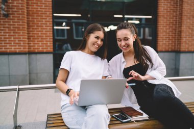Moda giysiler içinde gülen bayan öğrenci taşınabilir bilgisayarda sınıf arkadaşıyla komik videolar paylaşırken kampüs karşısında birlikte bankta dinleniyor.