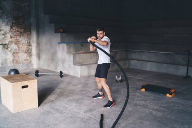 Spor kıyafetli, kaslı bir erkek sporcunun savaş halatını çevirmesi spor salonunda omuz kaslarını güçlendirme egzersizi yaparken kameraya bakarken viziteye giriyor.