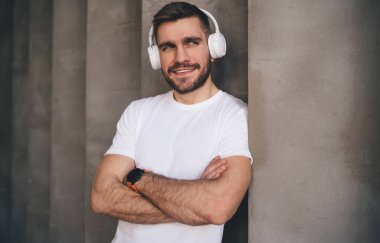 İyimser sakallı, beyaz t-shirt ve kulaklık takan, kollarını kavuşturmuş beton duvara yaslanmış modern şarkılar dinleyen erkek müzik aşığı.
