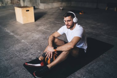 Spor kıyafetleri içinde yüksek açılı sakallı erkek sporcu mindere oturup, antrenmandan sonra kablosuz kulaklıkla müzik dinliyor.