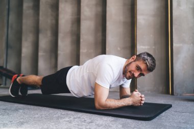 Beyaz tişörtlü güçlü bir erkek sporcu spor salonunda spor yaparken ve kameraya bakarken minder üzerinde tahta egzersizi yapıyor.