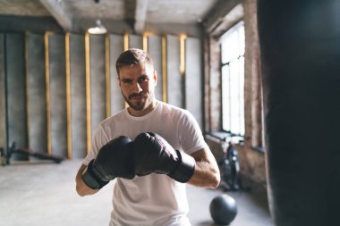 Boks eldivenli ciddi sakallı erkek boksör kameraya bakıyor ve spor salonunda egzersiz yaparken ön planda siyah torbayla yumruk pratiği yapıyor.