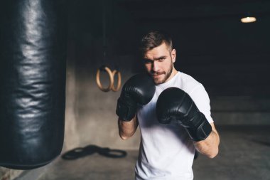 Boks eldivenli güçlü erkek boksör spor salonunda antrenman yaparken ön planda kum torbasına saldırmayı hedeflerken kameraya bakıyor.