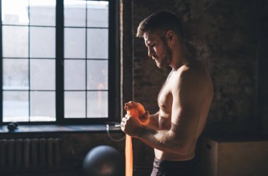 Pencere kenarında duran kaslı erkek sporcunun yan görünüşü ve spor salonunda boks yaparken el bağlarını ve eklemlerini dikkatle korumak için avuç içi ve baş parmaklarını sarmalamak.
