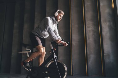 Spor giyim ve kablosuz kulaklık takan, bisiklet simülatörü eğitimi alan ve spor salonunda müzik dinleyen sakallı bir erkek sporcunun yan görüntüsü.