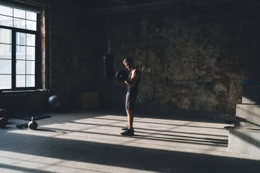 Güneşli bir günde pencerenin önünde duran sporcunun antrenman sırasında halterlerle kaslarını şişirmesi.