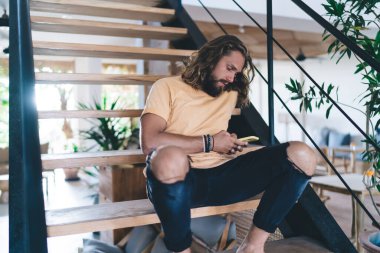 Şık kıyafetli, kıvırcık saçlı, cep telefonu kullanırken mesajlaşan ve modern evin merdivenlerinde dinlenen bir erkekten.