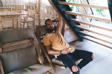 Yüksek açılı, sakallı bir erkek, düşünceli bir şekilde rahatlıyor ve merdivenin altında cep telefonuyla rahat bir kanepeye bakıyor.