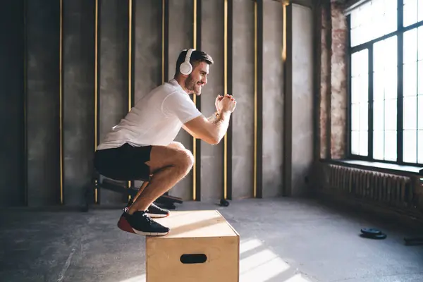 stock image Side view of positive male athlete in sportswear and headphones listening to music while practicing leg muscle exercise during functional training in modern loft style fitness club