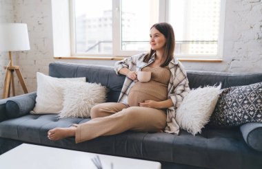 Yumuşak yastıklarıyla kanepede uzanan uzun boylu bir kadın kupayı karnına koyuyor ve doğum izninde harika vakit geçiriyor.