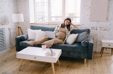 Stresli, genç, hamile, serbest çalışan, kanepede dinlenen ve çevrimiçi iş sırasında dizüstü bilgisayarını karıştıran ve evdeki rahat oturma odasında dinlenen bir kadın.