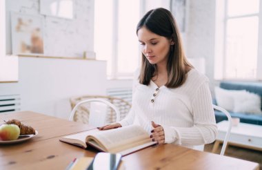 Yoğunlaşmış hamile kadın kitap okurken, hafta sonu evde hafif modern mutfakta meyve ve atıştırmalıklarla birlikte ahşap masada oturuyor.