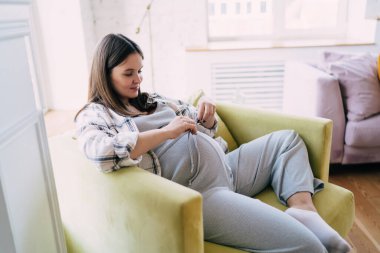 Uzun siyah saçlı, rahat koltukta dinlenen, uzun boylu, hamile bir annenin yan görüntüsü.