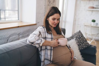 Positive female putting hand on belly feeling baby kicking while relaxing on couch in cozy lounge and drinking hot beverage clipart