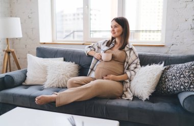 Yumuşak yastıklarıyla kanepede uzanan uzun boylu bir kadın kupayı karnına koyuyor ve doğum izninde harika vakit geçiriyor.