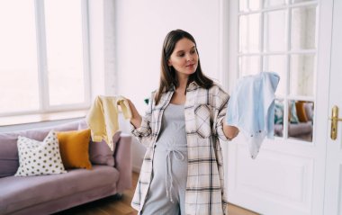 İki renkli bebek elbisesiyle gülümseyen ve ayakta duran çocuksu bir kadın. Evde rahat bir odada vakit geçiriyorlar.