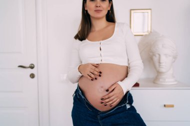 Beyaz uzun kollu bluz ve düğmesiz kot pantolon giyen kadın bebeği çıplak karnına hafifçe dokunurken büstü olan şifoniyerin yanında duruyor.