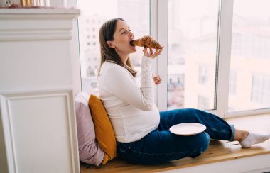 Hamilelik kıyafetleri giymiş, koca göbekli, sabah kahvaltı yapıp dinlenirken kruvasanın tadını çıkaran bir kadın bekliyor.