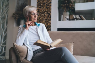Oturma odasında çenesine dokunarak ve ilginç kitaplar okuyarak kanepede dinlenirken gülümseyen ve başka tarafa bakan son sınıf öğrencisi pozitif bir kadın.